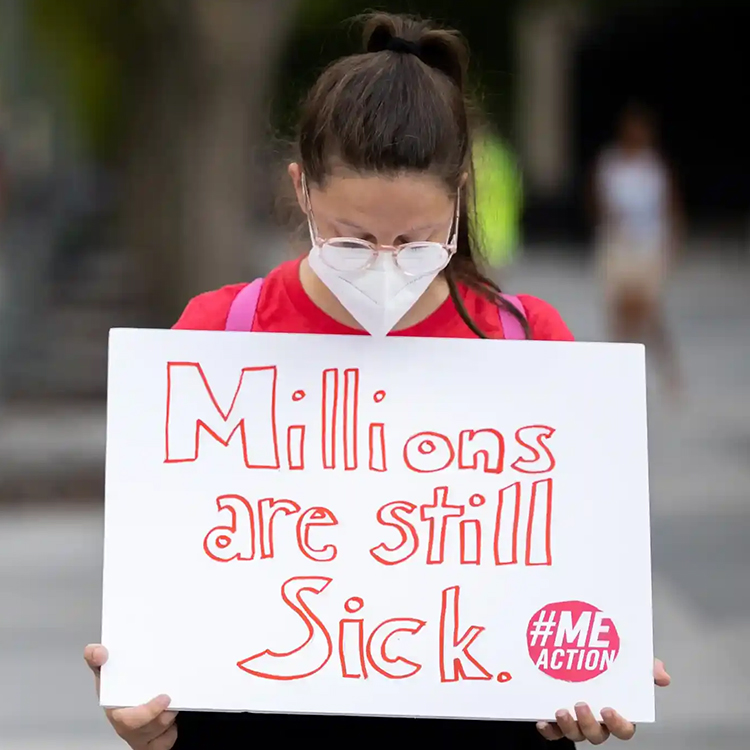 Woman wearing a mask with her head bowed, holding a sign that says, "Millions are still sick."