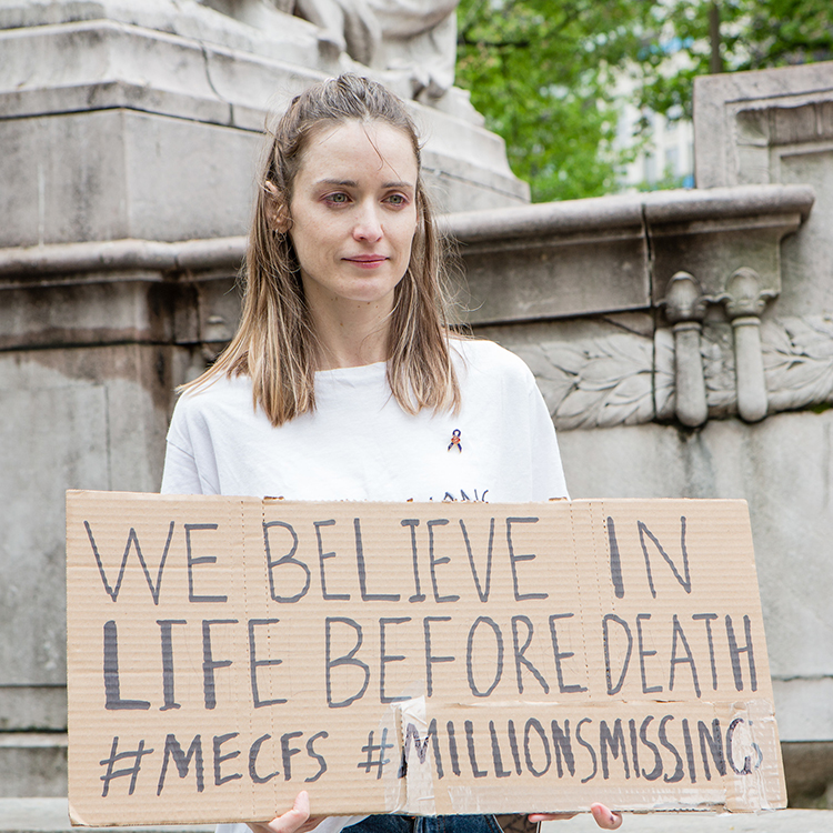 A woman holding a sign that says "We believe in life before death. #MECFS #MillionsMissing."