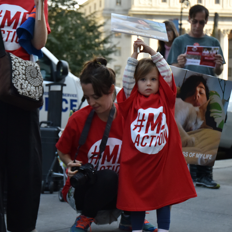 A little girl in a big #MEAction shirt. Her arms are outstretched, holding a sign above her head.