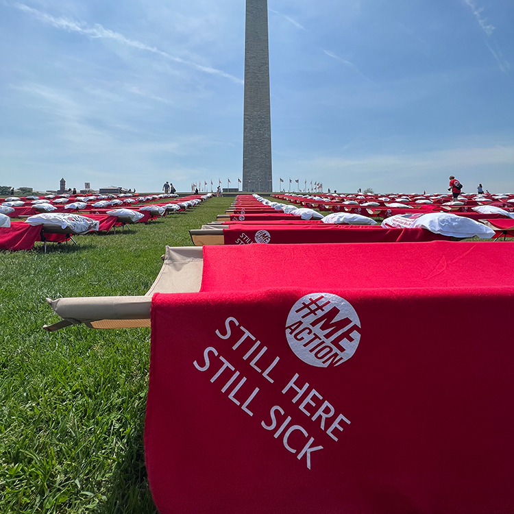 Closeup of a red cot that shows the #MEAction logo and the words "Still Here, Still Sick" on the lawn in front of the Washington Monument.